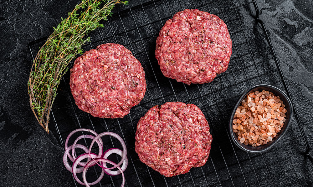 raw hamburger patties on wire rack with rosemary and onions at the side